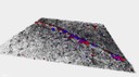 L'immagine mostra la ricostruzione tridimensionale di un frammnento di polietilene tereftalato (PET) dalla forma trapezoidale di colore grigio con una superficie irregolare su cui sono evidenziate mediante colorazione rossa e blu le diverse cellule batteriche presenti di forma sia allungata che tondeggiante.
