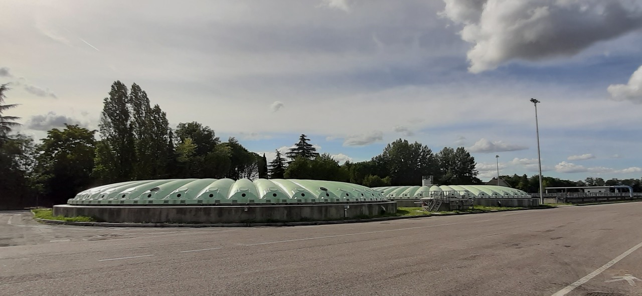 Un particolare dell’area dell’impianto di depurazione Hera di via Calcinaro a Cesena, con ii primo piano due vasche di sedimentazione primaria con copertura ondulata di colore verde. Sllo sfondo alberi e cielo parzialmente nuvoloso